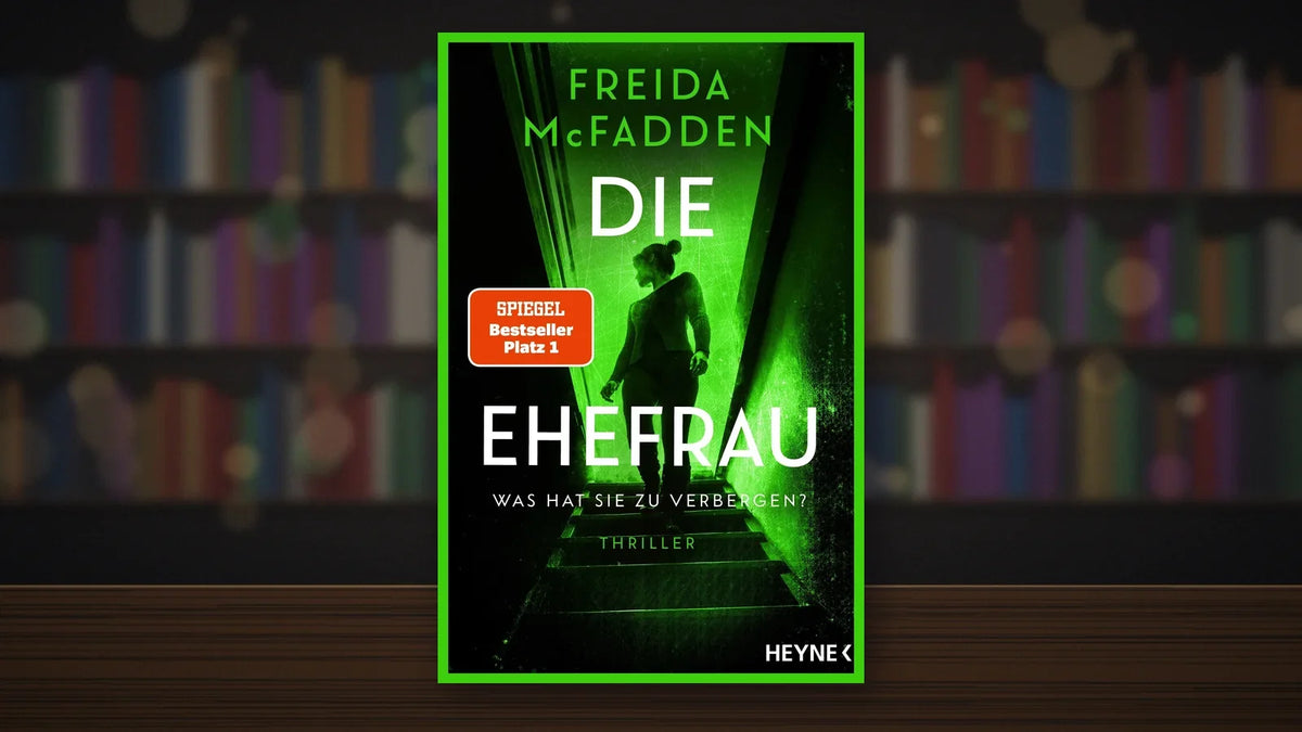 Buchcover von »Die Ehefrau« von Freida McFadden auf einem Holztisch vor unscharfen Bücherregalen in einer Buchhandlung